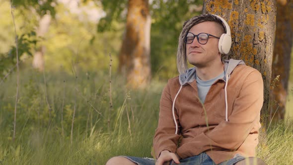 Young Relaxed Man in Glasses Headphones Sitting Near Tree in Green Grass Listening to Music on alt