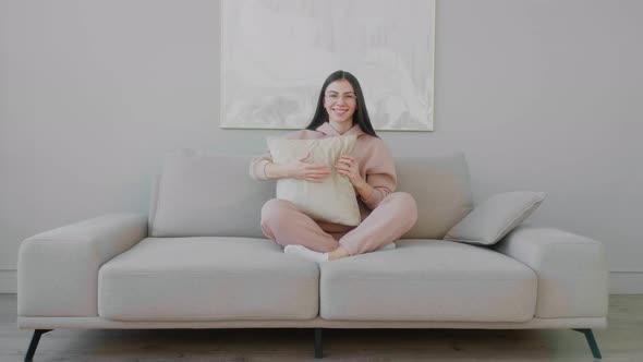 Young Caucasian woman with glasses sitting cross legged on the sofa and playfully tossing a pillow.