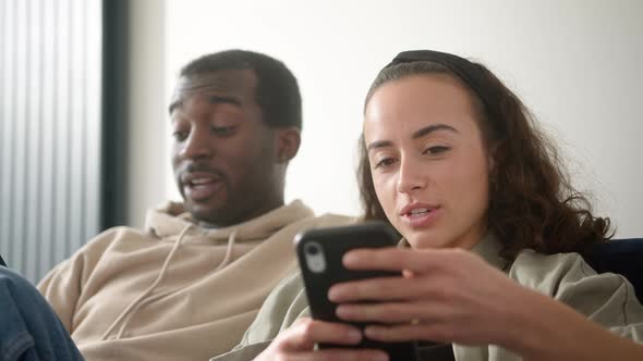 Young Smiling Couple Relaxing At Home On Sofa Checking Social Media On Mobile Phones Together
