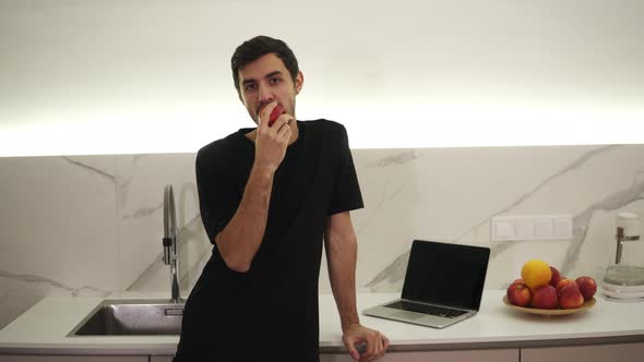Young Man Eating Red Apple at Home in the Kitchen alt