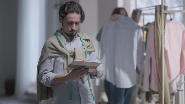 Young Concentrated Man Surfing Internet on Tablet As Woman Choosing Clothes From New Collection at alt