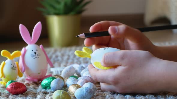A girl draws a pattern on an Easter egg in the form of a rabbit with Easter eggs in the background