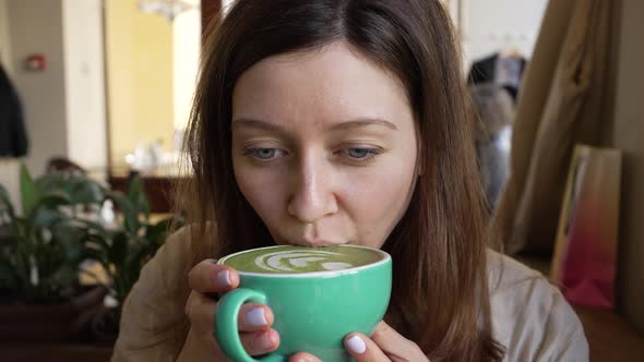 Young Woman in Cafe Takes Sip of Matcha Latte From Cup alt