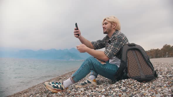 A Young Man in a Plaid Shirt Sits on the Shore and Takes Photo on His Phone of the Sea alt