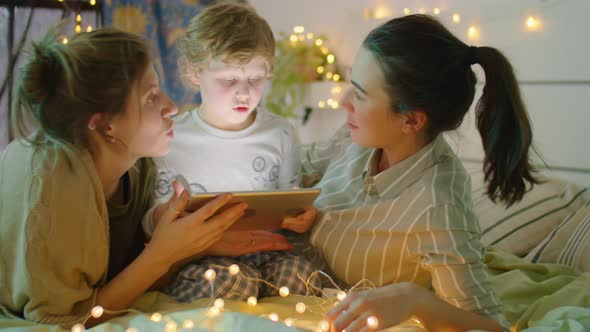 LGBTQ Family Using Tablet and Speaking on Bed with Garland alt