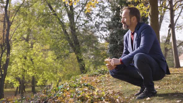 A Middleaged Caucasian Man Picks Up a Leaf Examines It and Looks Around in a Park in Fall