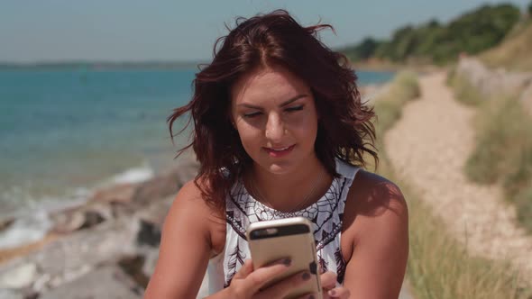 Portrait Of Young Attractive Woman By The Sea Using Her Phone To Make Online Purchase And Then Smili alt