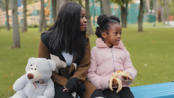 Portrait Young Mom and Daughter Sitting on Bench with Teddy Bear Child Eating Banana Woman Points alt