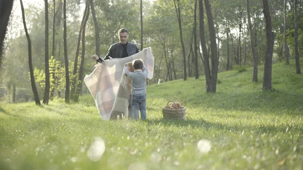 Wide Shot of Caucasian Young Father and Little Son Laying Blanket on Green Summer Meadow alt