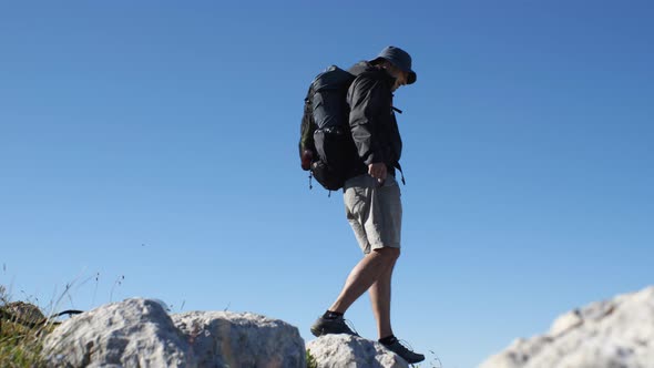 A man in shorts and a windbreaker with a backpack on his back jumps on rocks in the mountains agains alt