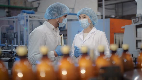 A Woman with a Young Colleague a Man in a White Coat and a Medical Mask at a Factory Near a Conveyor