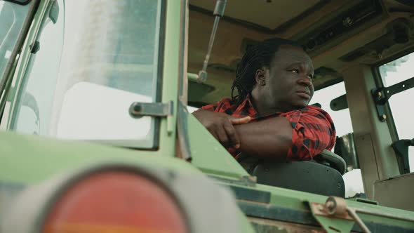Tired Young African Farmer Sitting in the Tractor Cabine and Looking Around alt