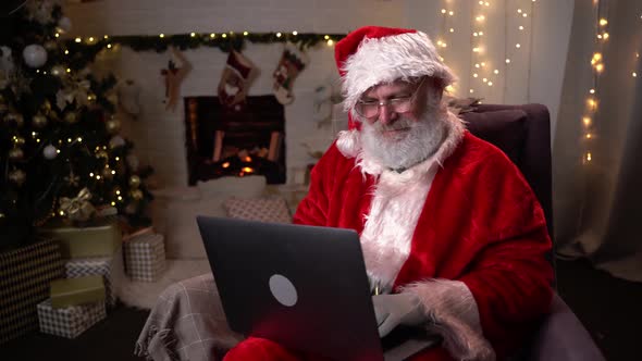 Modern Santa Claus. Cheerful Santa Claus Working on Laptop and Smiling While Sitting at His Chair