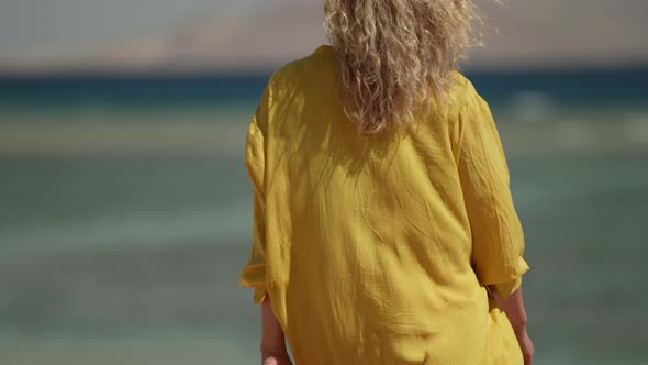 a Blonde in Sunglasses and a Yellow Fluttering Dress Walks Against the Background of a Blurred Sea alt