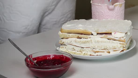 A Woman Smears A Biscuit Cake With Cream. Nearby Is A Container With Cherry Filling For The Cake. alt
