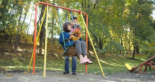 Bearded Husband Shakes a Rocking Chair with His Wife Holding a Bouquet of Autumn Leaves. Perky alt