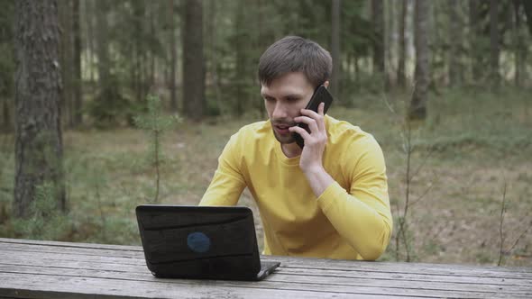 Dissatisfied Indignant Freelancer Sits in Forest Behind Laptop Calls on Phone alt