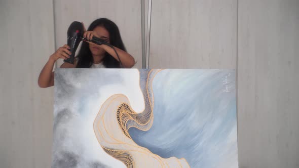 Female Artist Drying Top Side Of A Paint Canvas With A Hair Dryer In The Studio. medium shot alt