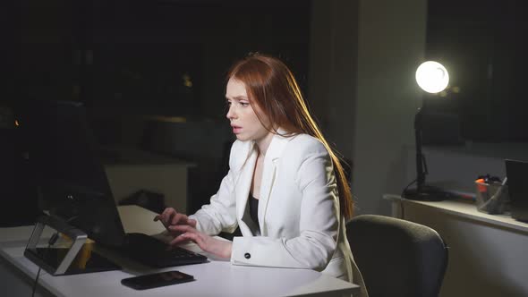 A Sleepy Redhaired Businesswoman Working at Night in the Office is Sitting at the Table Tired and alt