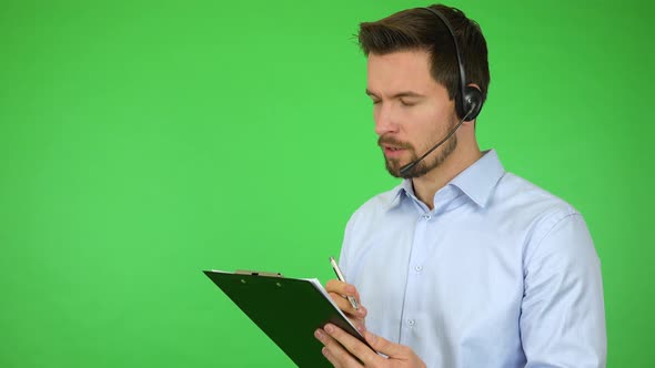 A Young Call Center Agent Talks To a Caller and Holds Notepad and a Pen, Then Smiles - Green Screen alt