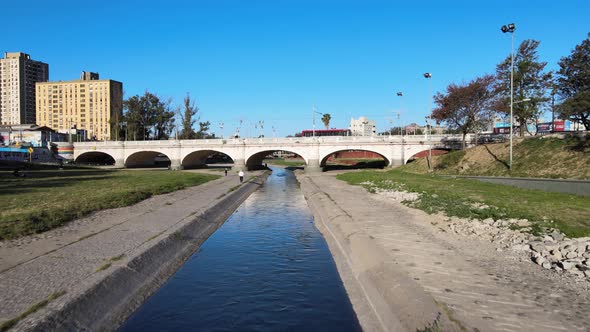 Low level aerial drone flying along narrow water canal toward Puente Centenario arch bridge, capturi alt