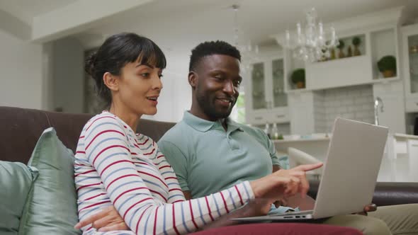 Happy diverse couple sitting on couch and using laptop in living room alt