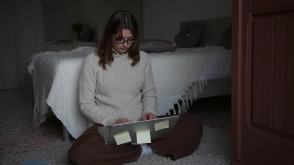 4k a girl with eyeglasses wearing sweater work with laptop, sitting on the floor in bedroom