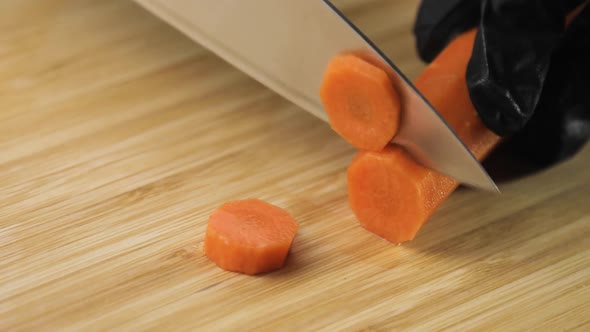 Cook's Hands in Black Gloves Cutting Fresh Carrot Into Slices on Wooden Board