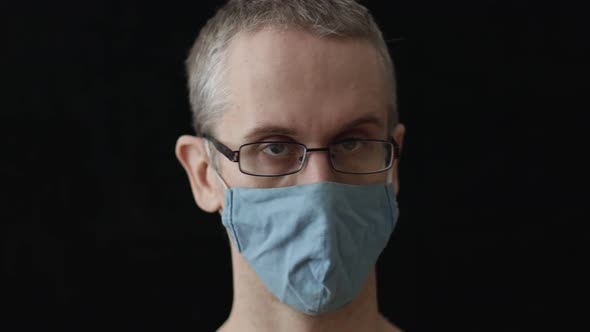 a Man in a Medical Mask and Glasses Shakes His Head Negatively on a Black Background