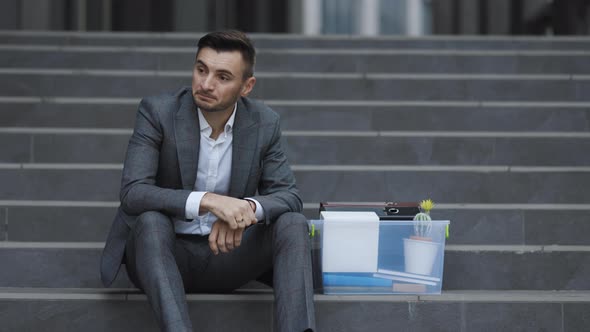 Businessman Sitting on Stairs Near Office Center With Box and Documents as Lost Job