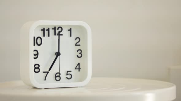 White alarm clock and glass of water on table in bedroom. Hand holding glass alt