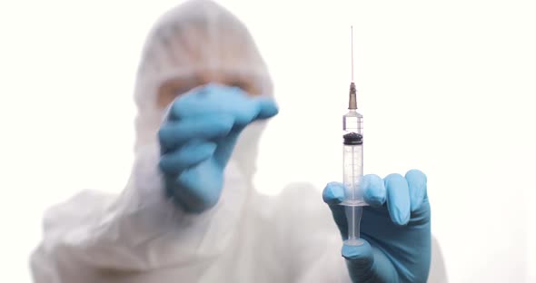 Doctor in a Protective Suit with a Syringe Looking to a Syringe with Vaccine