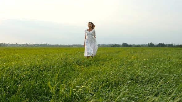 Beautiful Young Woman in White Dress Running on the Green Field. Wind Blowing Hair