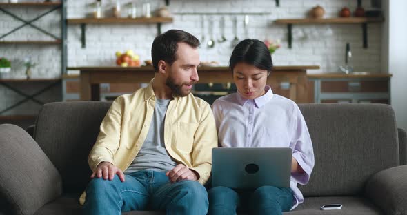 Happy Multiracial Young Couple Talking and Using Laptop Together Watching Video Sitting on Sofa in alt