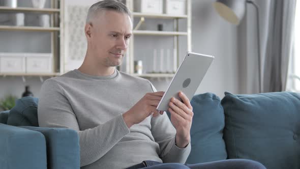 Gray Hair Man Browsing Internet on Tablet While Sitting on Couch alt