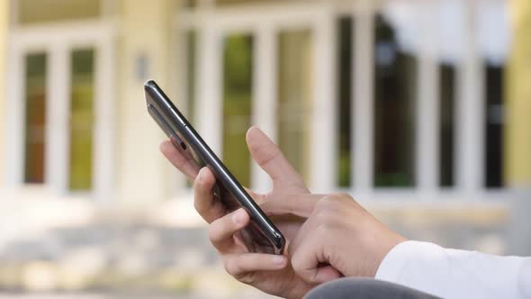 A Caucasian Teenage Boy Works on a Smartphone  Closeup on the Phone  a School in the Background