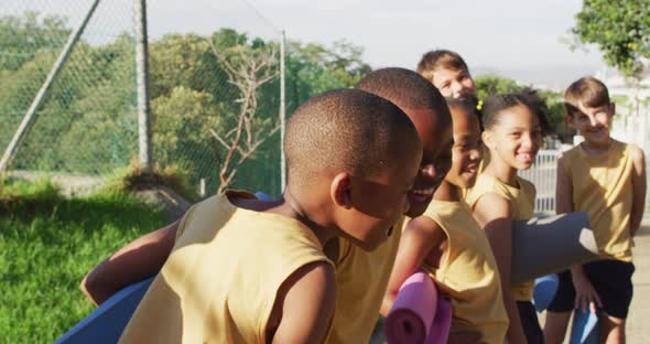 Diverse group of schoolchildren standing holding mats before yoga lesson outdoors alt