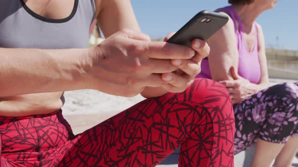 Caucasian women sitting down and woman using her phone on the beach and blue sky background alt