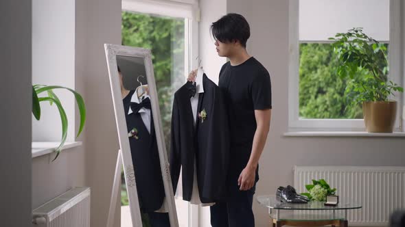 Young Groom Trying on Wedding Suit Looking in Mirror in Living Room alt