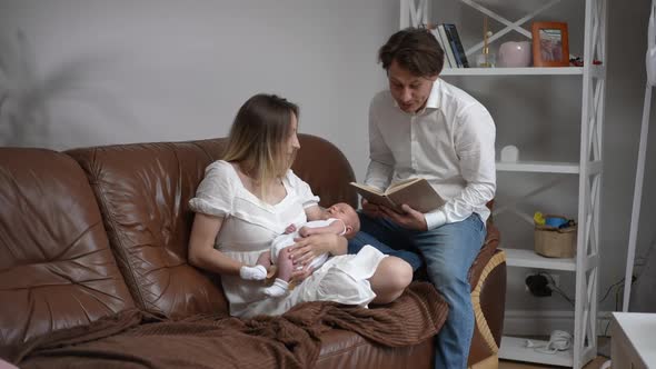 Caucasian Happy Father Reading Fairy Tales to Newborn Son in Hands of Smiling Woman Sitting on Cozy alt