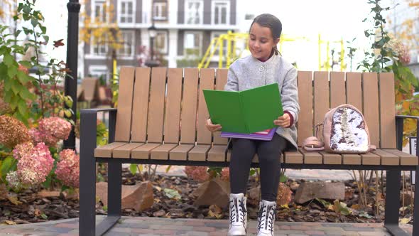 Schoolgirl Reading Book While Sitting on Bench in Park alt