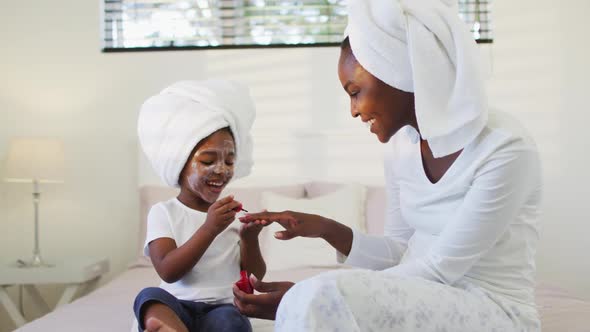 Happy african american mother and daughter sitting on bed and painting nails alt