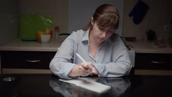 Woman Sits in the Kitchen at Home and Makes Plans Writes in a Notebook in the Evening
