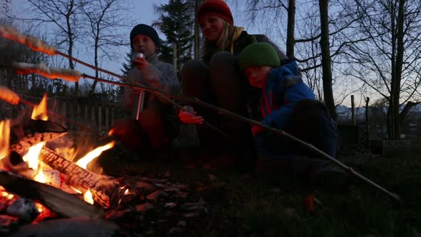 Two boys and their mother sitting around a bonfire with stick bread alt