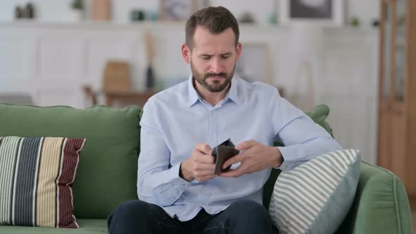 Broke Young Man Checking Empty Wallet on Sofa alt