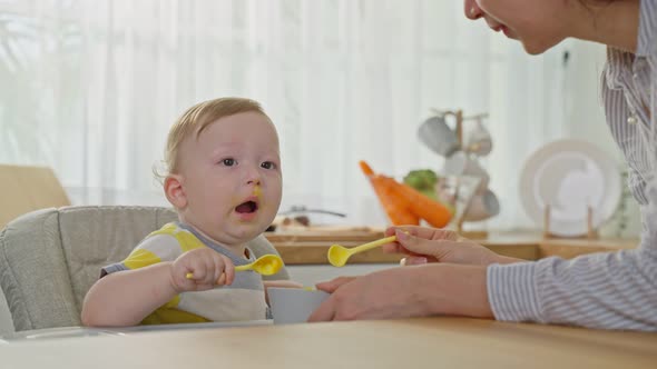 Caucasian beautiful mother take care and feeding healthy food to baby boy toddler in kitchen at home alt