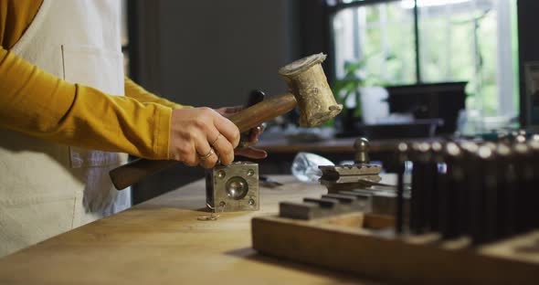 Midsection of caucasian female jeweller in workshop wearing apron, using hammer, making jewelry alt