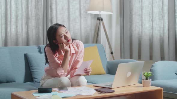 Asian Woman With A Laptop Looking At The Bill And Being Sad While Sitting On Sofa At Home alt