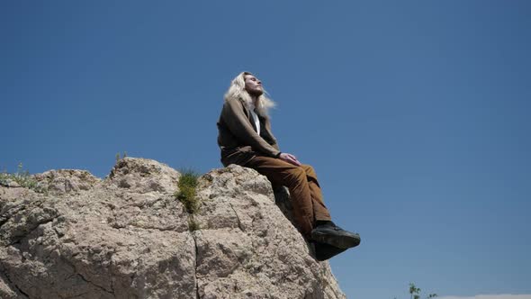 A Young Hipster Guy with Long Blond Hair Sits on the Edge of a Cliff