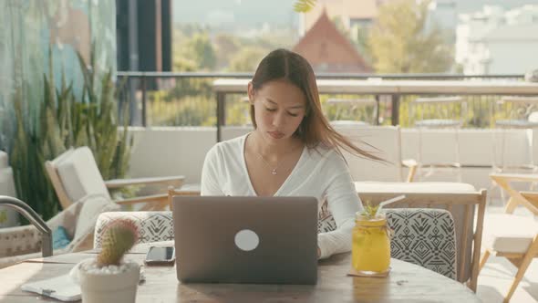 A Young Asian Girl Works at Her Laptop in a Rooftop Cafe in the City alt